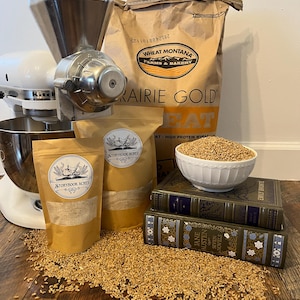 May include: A close-up of a grain mill with two bags of grain labeled "Storybook Acres" and a bag of "Prairie Gold" wheat. The grain mill is sitting on a wooden surface with a bowl of grain and a stack of books.