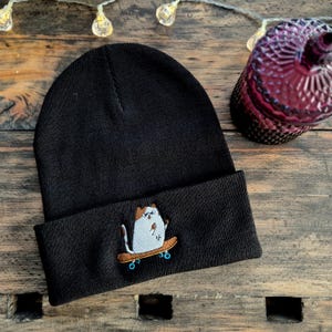 Könnte beinhalten: Schwarze Mütze mit einem gestickten weißen Kater, der auf einem Skateboard fährt. Der Kater trägt ein braunes Hemd und hat einen schwarzen Schwanz.