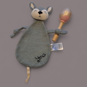 Può includere: Un giocattolo di peluche grigio a forma di topo con una faccia bianca e occhi neri. Il giocattolo ha una lunga coda e tiene un sonaglio di legno con una parte superiore rosa e dorata. Il giocattolo è ricamato con il nome "Yma".
