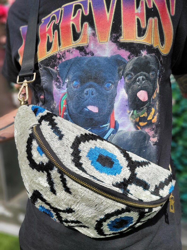 May include: A crescent-shaped bum bag featuring a textured velvet fabric with a repeating abstract eye pattern in shades of cream, black, and bright blue. The bag includes a black zip closure with gold-toned hardware and an adjustable black strap.