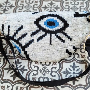 May include: A crescent-shaped bum bag featuring a white textured surface with a prominent black and blue evil eye graphic. The bag includes a black adjustable strap attached with gold-toned metal hardware clips on both sides of the main body.