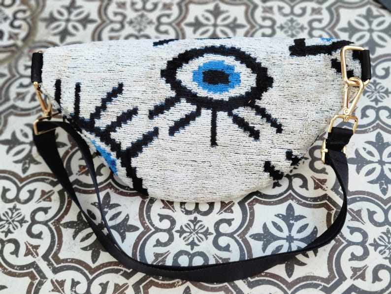 May include: A crescent-shaped bum bag featuring a white textured surface with a prominent black and blue evil eye graphic. The bag includes a black adjustable strap attached with gold-toned metal hardware clips on both sides of the main body.