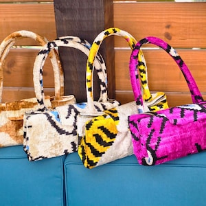 May include: Four rectangular handbags with single top handles, each featuring a distinct velvet ikat pattern. The bags are displayed in a row, showcasing color variations of tan, cream with blue accents, yellow with black stripes, and vibrant magenta.
