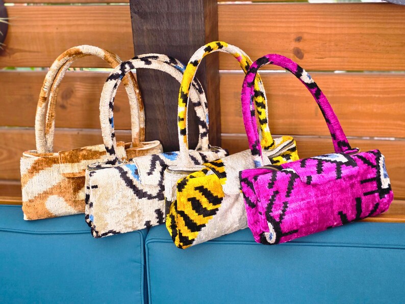 May include: Four rectangular handbags with single top handles, each featuring a distinct velvet ikat pattern. The bags are displayed in a row, showcasing color variations of tan, cream with blue accents, yellow with black stripes, and vibrant magenta.