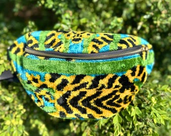 May include: A bum bag featuring a vibrant velvet texture with a bold, abstract pattern in shades of bright yellow, turquoise blue, and black. The bag includes a horizontal green velvet stripe across the front and a dark metal zipper closure.