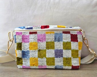May include: A rectangular crossbody bag featuring a multi-colored checkered pattern in shades of yellow, red, blue, pink, and olive green. The bag has a white zipper closure, gold-tone metal hardware, and a wide, solid cream-colored fabric strap.