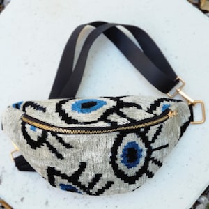 May include: A crescent-shaped bum bag featuring a textured silver-grey fabric with a repeating black and blue eye pattern. The bag includes a gold-toned metal zip closure and an adjustable black strap with gold-coloured metal hardware connectors.