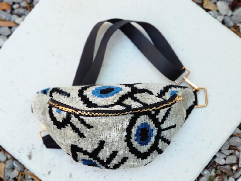 May include: A crescent-shaped bum bag featuring a textured silver-grey fabric with a repeating black and blue eye pattern. The bag includes a gold-toned metal zip closure and an adjustable black strap with gold-coloured metal hardware connectors.