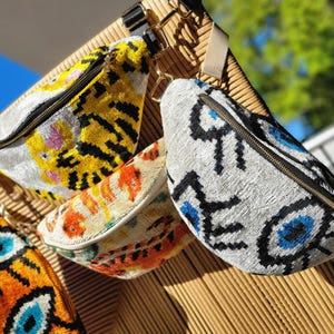 May include: Three bum bags featuring textured fabric with distinct patterns. One displays a yellow and black tiger motif, another shows an orange and cream abstract design, and the third features a white background with blue and black eye symbols.