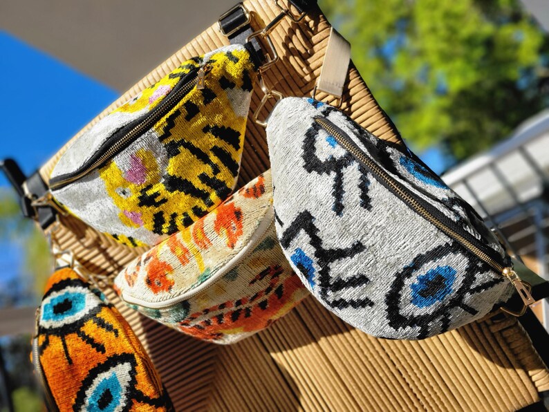 May include: Three bum bags featuring textured fabric with distinct patterns. One displays a yellow and black tiger motif, another shows an orange and cream abstract design, and the third features a white background with blue and black eye symbols.