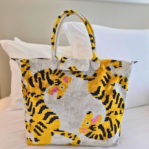May include: A tote bag made of a textured, light gray fabric featuring a repeating pattern of two yellow tigers with black stripes and pink accents. The bag has two matching handles and a top zipper closure, with the tiger design extending onto the handles.