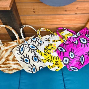 May include: Four tote bags with two handles each. The first bag has a brown and white tiger stripe pattern. The second bag is cream with a blue and black evil eye print. The third bag is white with a yellow tiger graphic. The fourth bag is bright pink with a blue and black evil eye print.
