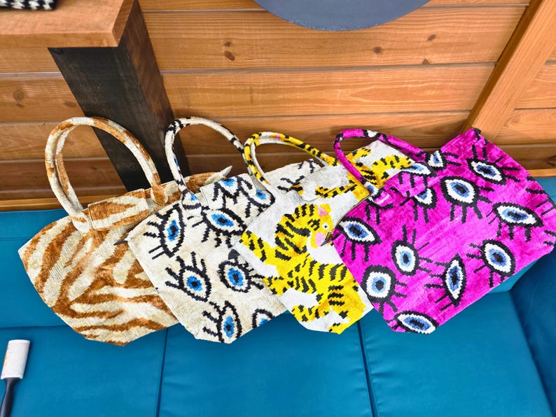 May include: Four tote bags with two handles each. The first bag has a brown and white tiger stripe pattern. The second bag is cream with a blue and black evil eye print. The third bag is white with a yellow tiger graphic. The fourth bag is bright pink with a blue and black evil eye print.