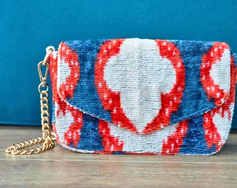 May include: A rectangular clutch bag made of textured velvet fabric featuring a repeating ikat pattern in shades of deep blue, bright red, and off-white. The bag includes a gold-tone metal chain strap attached with a lobster clasp on the side.