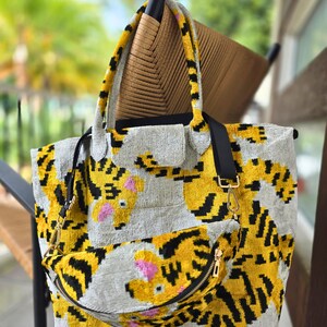 May include: A tote bag and a matching fanny pack featuring a yellow, black, and gray tiger stripe pattern with pink accents. Both items have a textured, velvet-like surface, black fabric straps, and gold-toned metal hardware clips and zippers.