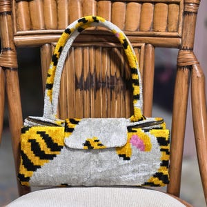 May include: A rectangular handbag featuring a velvet-like texture with a pattern of yellow, black, and silver-grey stripes. The bag includes a single arched handle, a central flap closure, and a zipper along the top edge with a small pink accent.