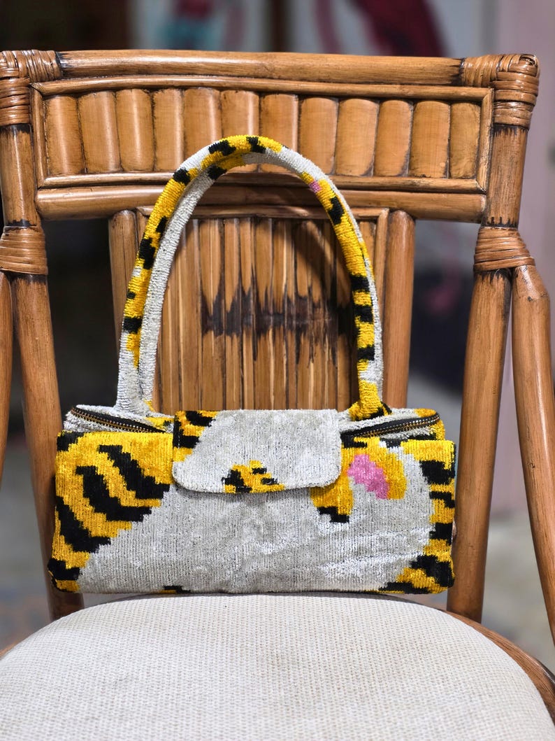 May include: A rectangular handbag featuring a velvet-like texture with a pattern of yellow, black, and silver-grey stripes. The bag includes a single arched handle, a central flap closure, and a zipper along the top edge with a small pink accent.