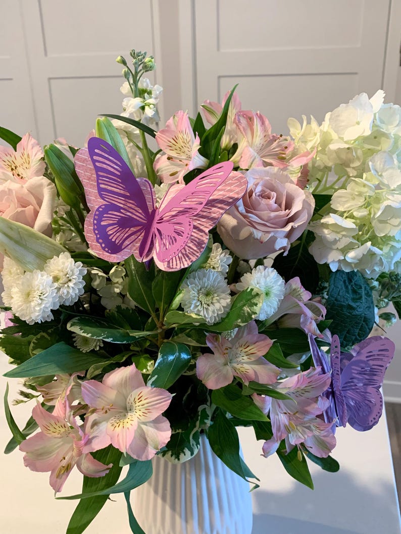 以下が含まれることがあります： 白い花瓶にピンクと白の花束と紫色の紙の蝶々
