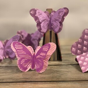 以下が含まれることがあります： 紫色の蝶の形をした紙製のしおり。しおりは重ねられており、3D効果があります。木製の表面に配置されています。