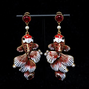 May include: A pair of red and white goldfish earrings with gold accents. Each earring features a red gem at the top, a pearl, and a detailed fish design with red, white, and gold enamel. The earrings are displayed against a black background.