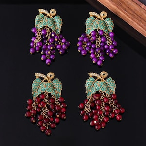 May include: Four pairs of grape-themed earrings. Each earring features a gold-colored leaf design with teal-colored accents and a cluster of purple or red beads. The earrings are on a black surface.