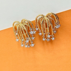 Puede incluir: Un par de pendientes dorados con un diseño texturizado. Cada pendiente presenta una estructura en forma de abanico con detalles colgantes en forma de estrella. El fondo es naranja.