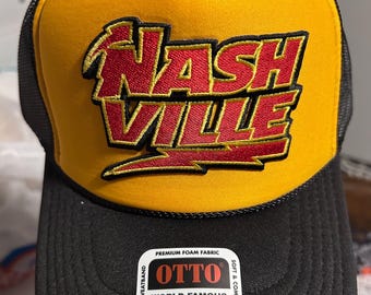 May include: A black and yellow trucker hat with a red and yellow embroidered "Nashville" logo. The hat has a black mesh back and a black brim. The hat has a label that reads "Otto World Famous Foam Front OSFM Trucker Durable Snap Closure".