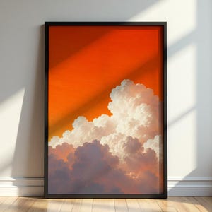 Op de afbeelding: Een ingelijste kunstdruk met een levendige oranje lucht en grote, pluizige witte en grijze wolken. Het kunstwerk wordt gepresenteerd in een zwarte lijst en leunt tegen een witte muur met een houten vloer.