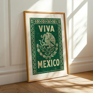 A framed green print with a distressed texture, featuring a white eagle on a cactus with a snake in its beak, and the words "VIVA MEXICO" above and below, leans against a white wall on a light wood floor.