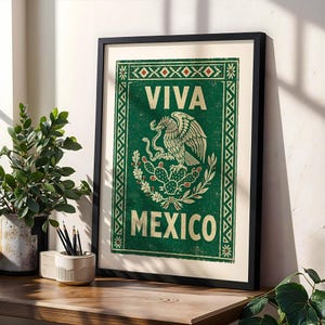 A framed green print with a distressed texture, featuring a white eagle on a cactus with a snake in its beak, and the words "VIVA MEXICO" above and below, leans against a wall on a wooden desk next to a potted plant and pencils.
