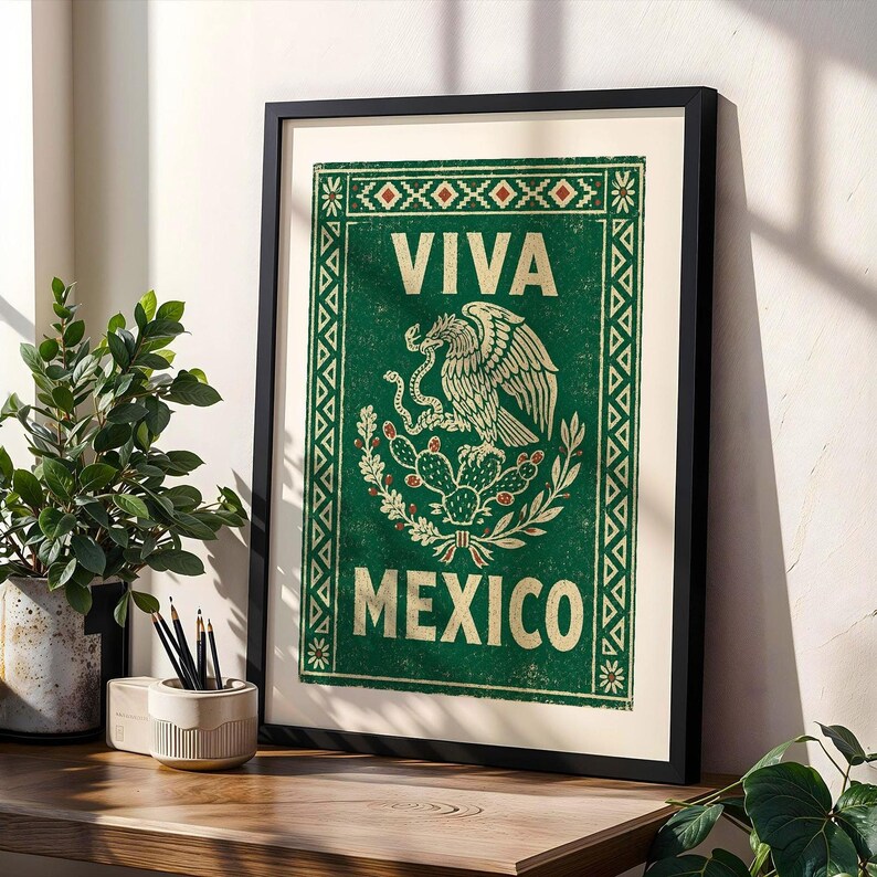 A framed green print with a distressed texture, featuring a white eagle on a cactus with a snake in its beak, and the words "VIVA MEXICO" above and below, leans against a wall on a wooden desk next to a potted plant and pencils.