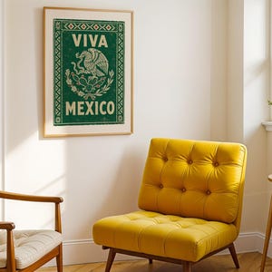 A framed green print with a distressed texture, featuring a white eagle on a cactus with a snake in its beak, and the words "VIVA MEXICO" above and below, hangs on a white wall above a yellow armchair in a living room setting.