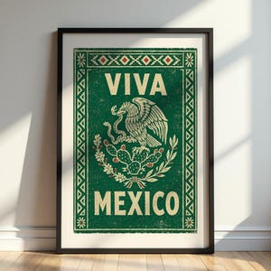 A framed green print with a distressed texture, featuring a white eagle on a cactus with a snake in its beak, and the words "VIVA MEXICO" above and below, leans against a white wall on a dark wood floor.