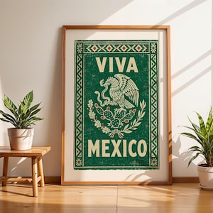 A framed green print with a distressed texture, featuring a white eagle on a cactus with a snake in its beak, and the words "VIVA MEXICO" above and below, stands on a light wood floor between two potted plants.