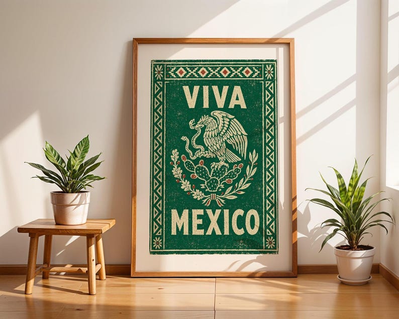A framed green print with a distressed texture, featuring a white eagle on a cactus with a snake in its beak, and the words "VIVA MEXICO" above and below, stands on a light wood floor between two potted plants.