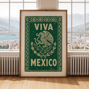 A framed green print with a distressed texture, featuring a white eagle on a cactus with a snake in its beak, and the words "VIVA MEXICO" above and below, stands on a light wood floor in front of a window overlooking a city and mountains.
