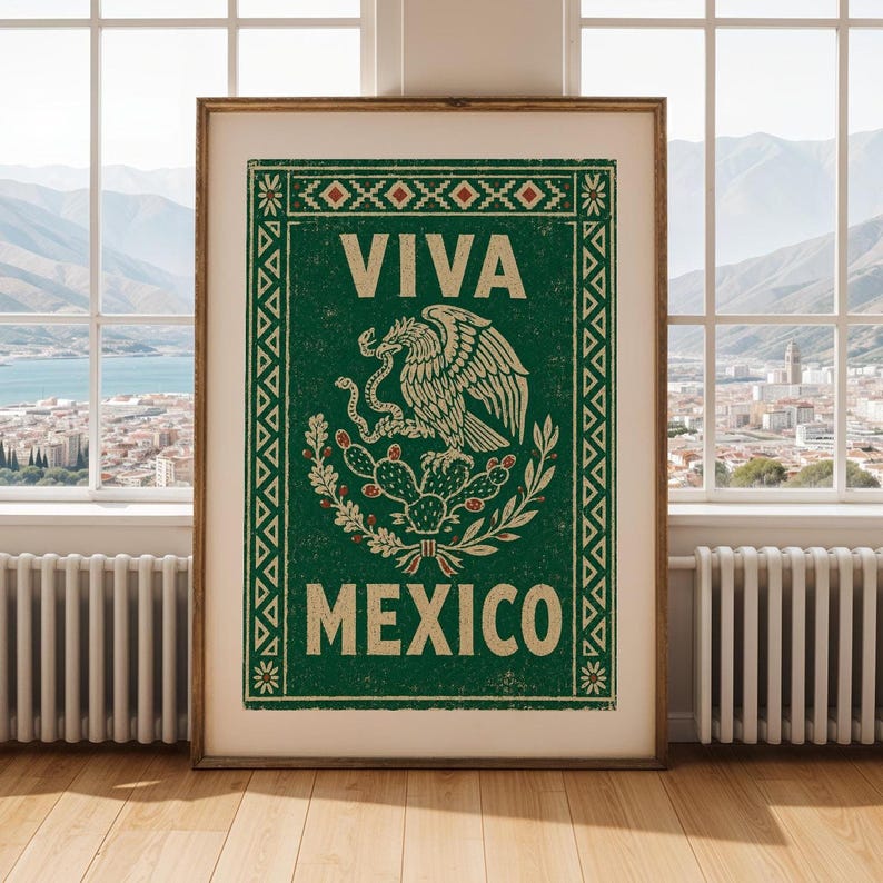 A framed green print with a distressed texture, featuring a white eagle on a cactus with a snake in its beak, and the words "VIVA MEXICO" above and below, stands on a light wood floor in front of a window overlooking a city and mountains.