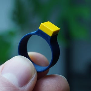 Könnte beinhalten: Ein blauer Silikonring mit einem gelben Quadrat oben.