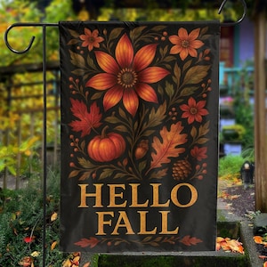 Op de afbeelding: Een zwarte tuinvaandel met een bloemen- en herfstontwerp. De vlag toont oranje bloemen, bladeren, een pompoen en de woorden "HELLO FALL" in gouden letters. De vlag wordt getoond op een zwarte metalen standaard.