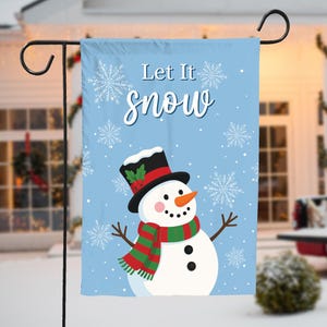 May include: A light blue garden flag featuring a snowman with a black top hat, red and green scarf, and an orange carrot nose. The flag reads "Let It Snow" with snowflakes in the background. The flag is displayed on a black metal stand.
