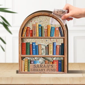May include: A wooden bookshelf-shaped money bank with a floral interior and a wooden sign that reads "Sarah's Library Fund." The bank is filled with colorful, decorative book cutouts and cash. A hand is inserting a bill into the slot.