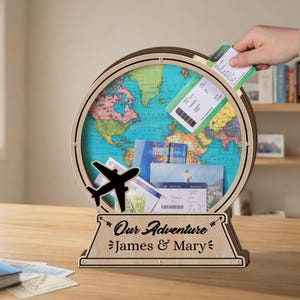 May include: A wooden globe-shaped shadow box with a world map background. A black airplane cutout is on the front. The base reads "Our Adventure" and "James & Mary". Tickets are being placed inside the globe.
