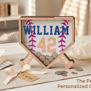 Puede incluir: Un banco de dinero con tem&aacute;tica de b&eacute;isbol con el nombre "WILLIAM" y el n&uacute;mero "42" en azul y melocot&oacute;n. El banco tiene forma de plato de home y contiene monedas. Una etiqueta de regalo con un lazo est&aacute; en primer plano.
