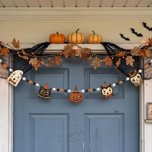 May include: A Halloween garland featuring felt pom-poms in white, orange, and black, interspersed with vintage-style cutouts of a ghost, a black cat, a jack-o'-lantern, a devil, and a skull, all strung across a blue door with autumn decor.