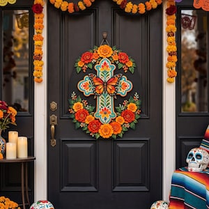 May include: A decorative wall hanging featuring a central cross adorned with a monarch butterfly and vibrant orange and red marigold flowers. The cross displays intricate blue, red, and yellow patterns, suspended on a black door with floral garlands.