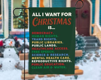 Può includere: Una bandiera da giardino verde scuro con la scritta "ALL I WANT FOR CHRISTMAS IS..." in bianco e rosso. La bandiera elenca vari diritti e valori in diversi colori, tra cui democrazia, diritti umani e accesso all'assistenza sanitaria.