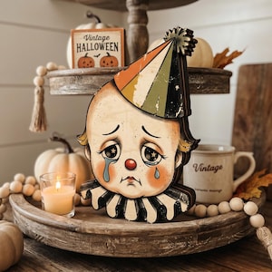 May include: A wooden cutout of a sad clown face with a red nose, blue teardrops, and large eyes with star pupils. The clown wears a multi-coloured party hat in orange, green, and black, and a black and white ruffled collar around the neck.