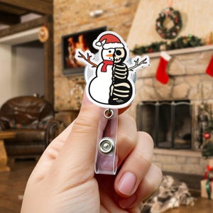 May include: A novelty badge reel featuring a snowman design, half of which is a skeleton. The snowman wears a red scarf and hat. The badge reel is clear plastic with a metal clip and button.