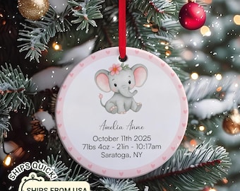 May include: A round, personalized Christmas ornament featuring a baby elephant illustration. The ornament has a pink border with hearts and includes the name "Amelia Anne", birthdate, weight, and location. A red ribbon is attached for hanging.