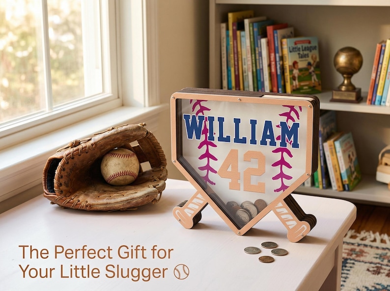 Puede incluir: Un banco de dinero con tem&aacute;tica de b&eacute;isbol en forma de plato de home, con el nombre "WILLIAM" y el n&uacute;mero "42". Un guante y una pelota de b&eacute;isbol est&aacute;n junto al banco. El banco tiene un marco de madera y est&aacute; lleno de monedas.